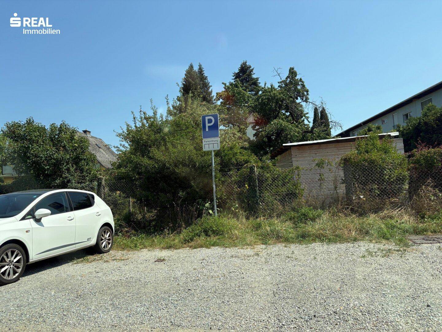 Kiesparkplatz mit Parkschild und angrenzendem, bewachsenem Grundstücksbereich.