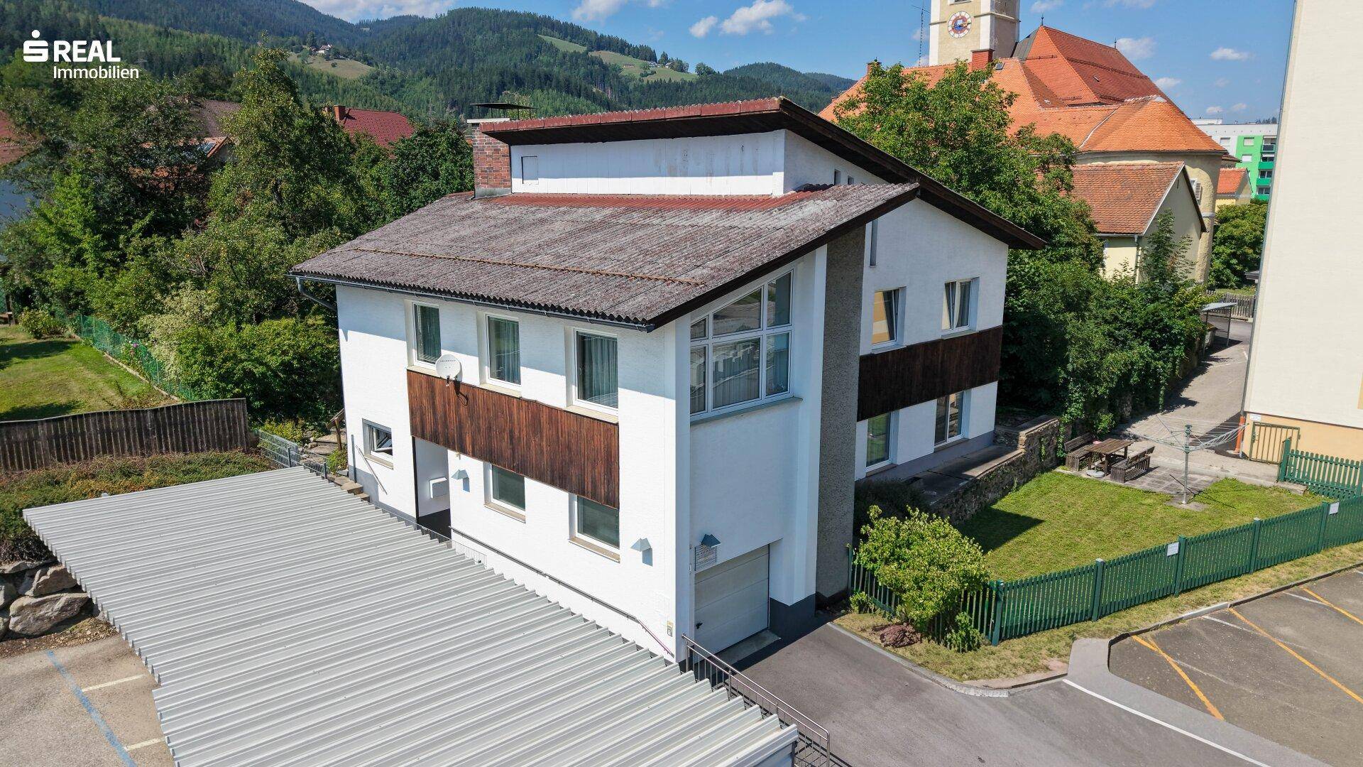 Schrägansicht des Hauses mit Carport und umliegender grüner Landschaft unter blauem Himmel.