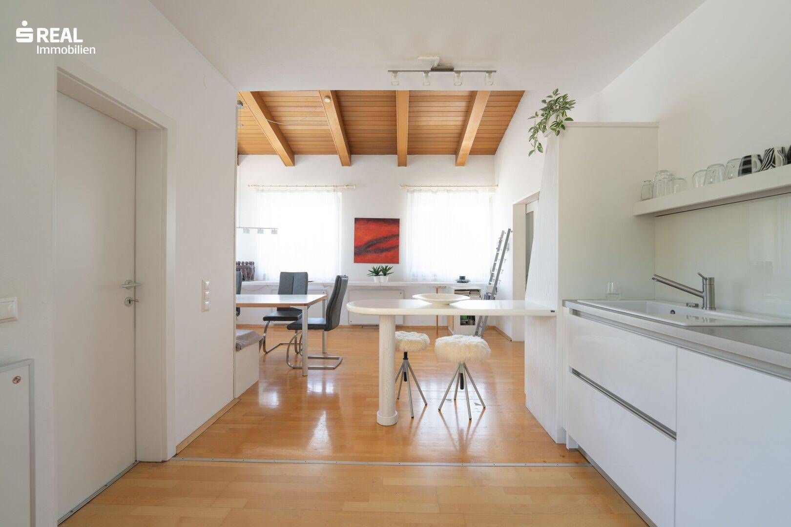 Helle Küche mit weißer Einbauküche, Frühstücksbar und Blick in den Essbereich mit Holzbalkendecke.