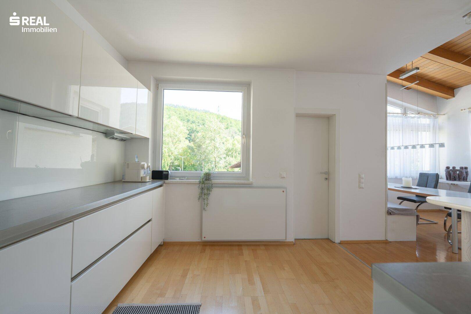 Moderne weiße Küche mit Fenster und Blick ins Grüne, angrenzend an den Essbereich.