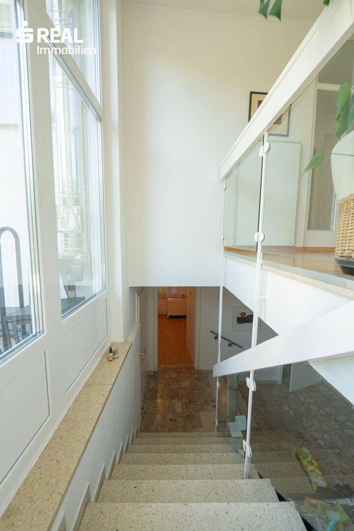 Treppenhaus mit großen Fenstern und hellem Steinboden, das die verschiedenen Ebenen verbindet.
