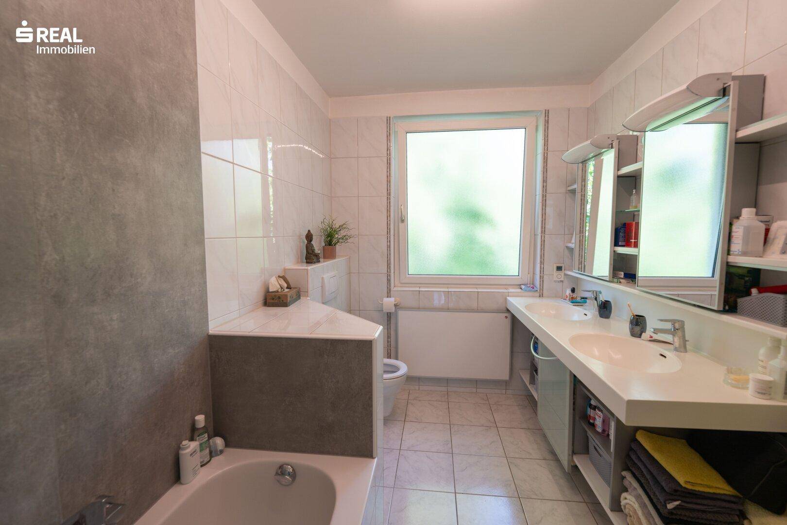 Geräumiges Badezimmer mit Doppelwaschbecken, Badewanne und Fenster, komplett gefliest.