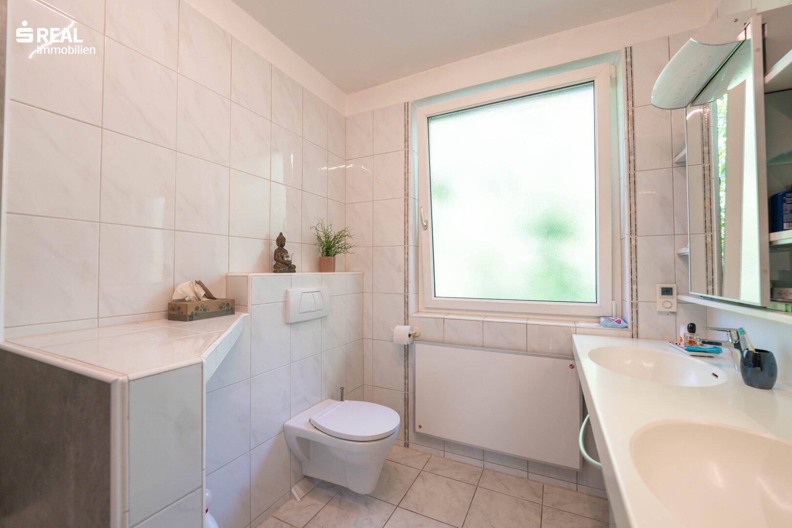 Helles Badezimmer mit WC, Doppelwaschbecken und Fenster, modern gefliest.