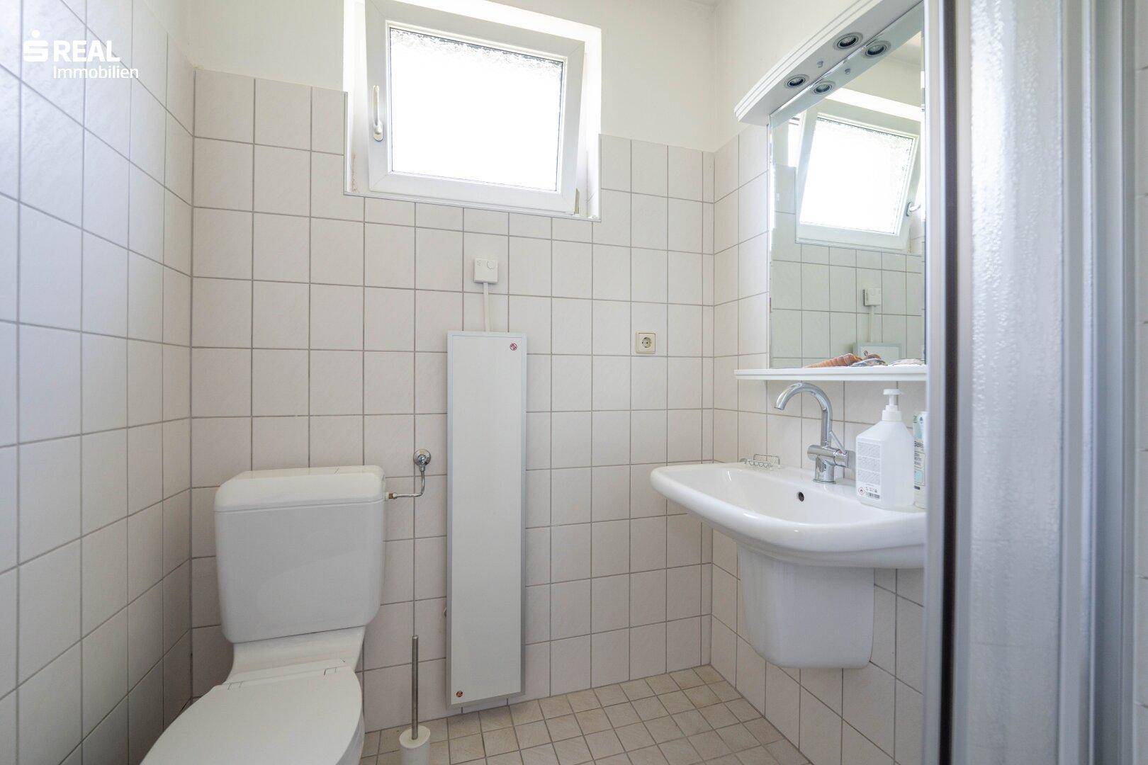 Badezimmer mit Dusche, WC und Waschbecken, hell gefliest und mit Fenster.