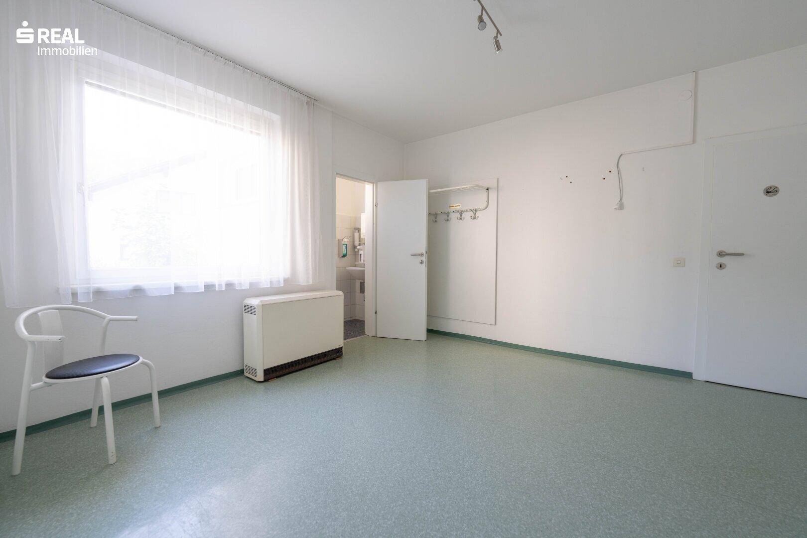 Leerer Bürobereich mit großem Fenster, Heizkörper und Zugang zu den Sanitäranlagen.