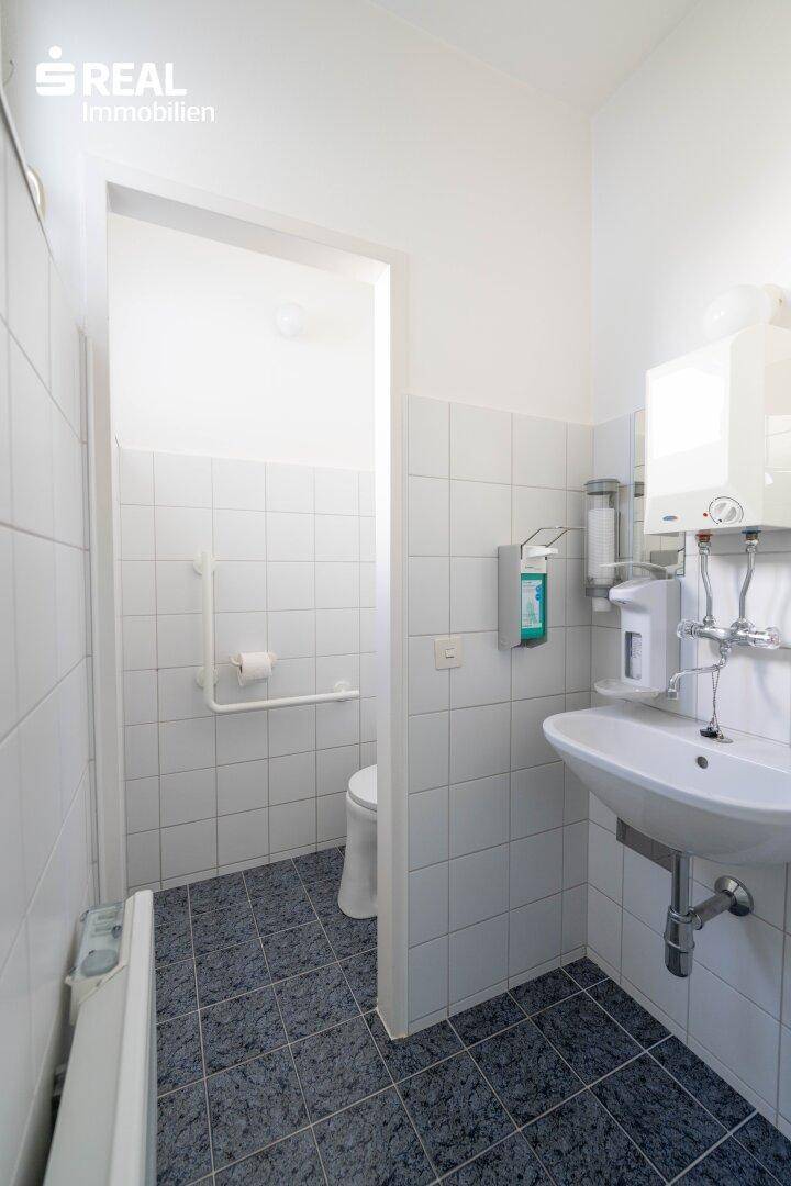 Funktionale Toilette mit weißen Fliesen, Waschbecken und Haltegriffen für Barrierefreiheit.