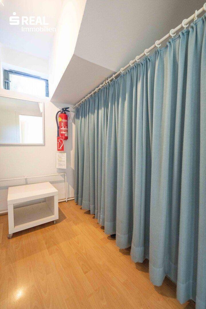 Flur oder Umkleidebereich mit hellblauen Vorhängen, Holzfußboden und einem kleinen Fenster.