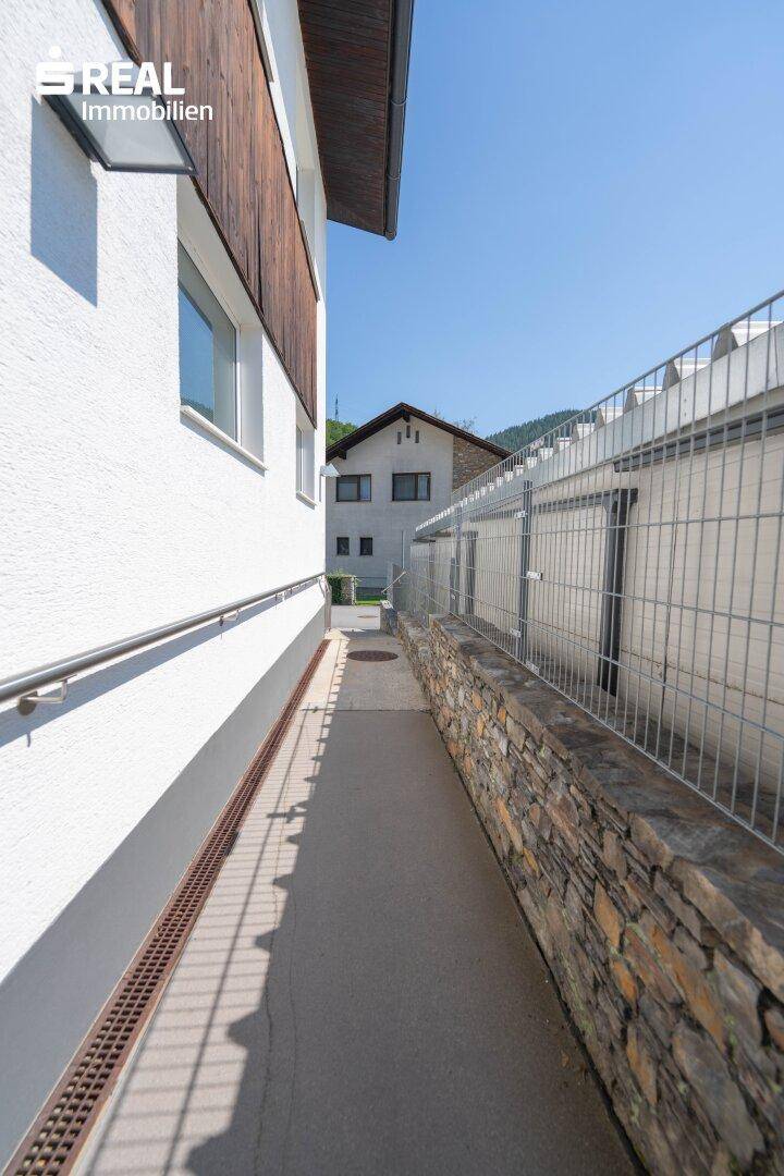 Schmaler Zugangsweg mit Handlauf entlang der Hauswand, führt zu einem Nebengebäude.