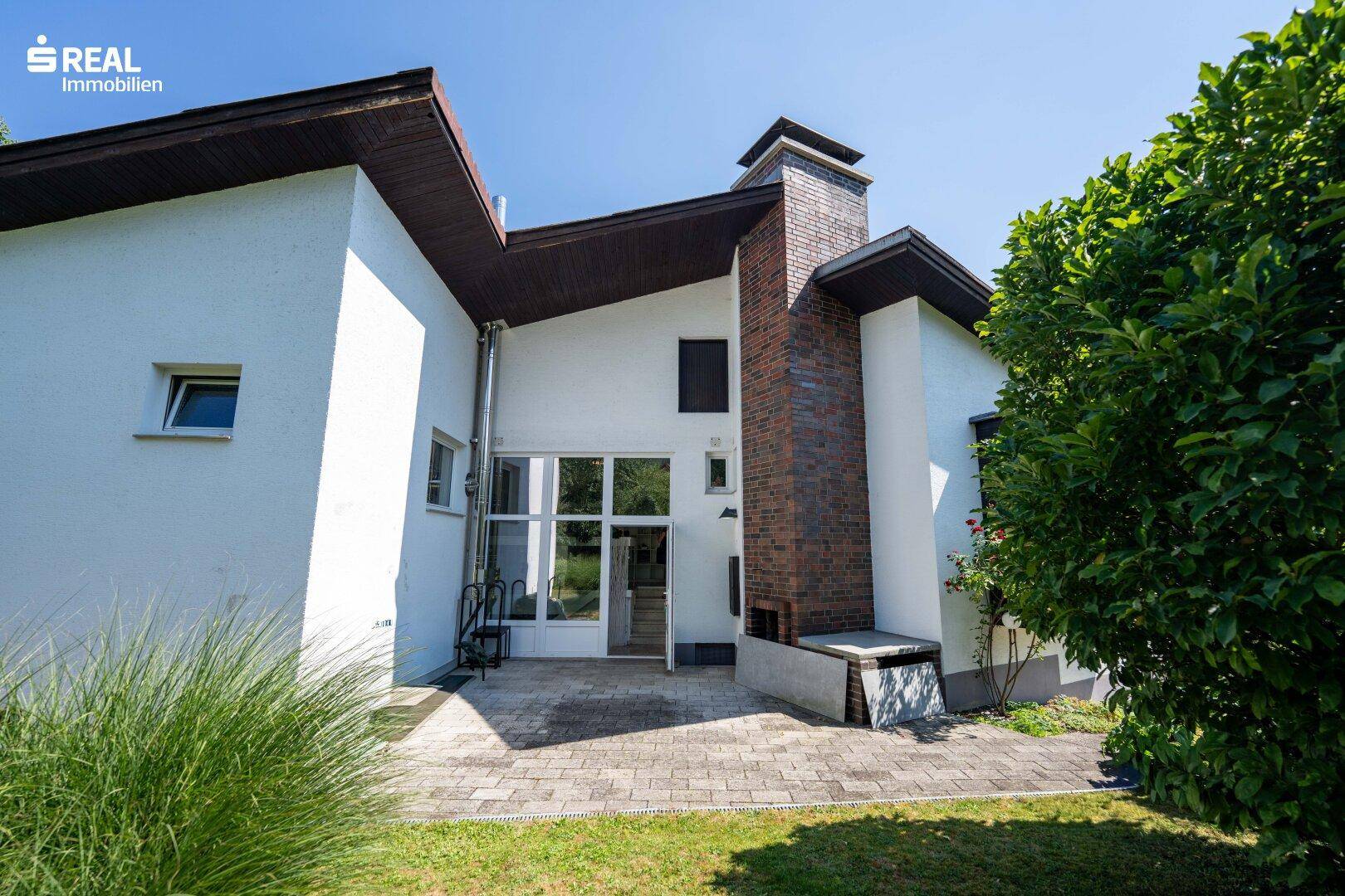 Sonniger Terrassenbereich mit gemauertem Grill und Zugang zum Haus, umgeben von Grün.