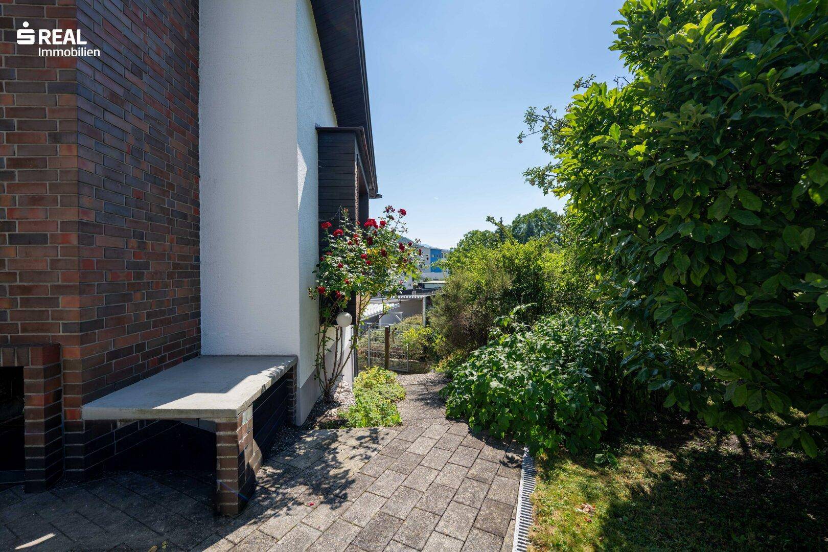 Gepflasterter Weg entlang des Hauses mit üppiger Bepflanzung und Blick in die Ferne.