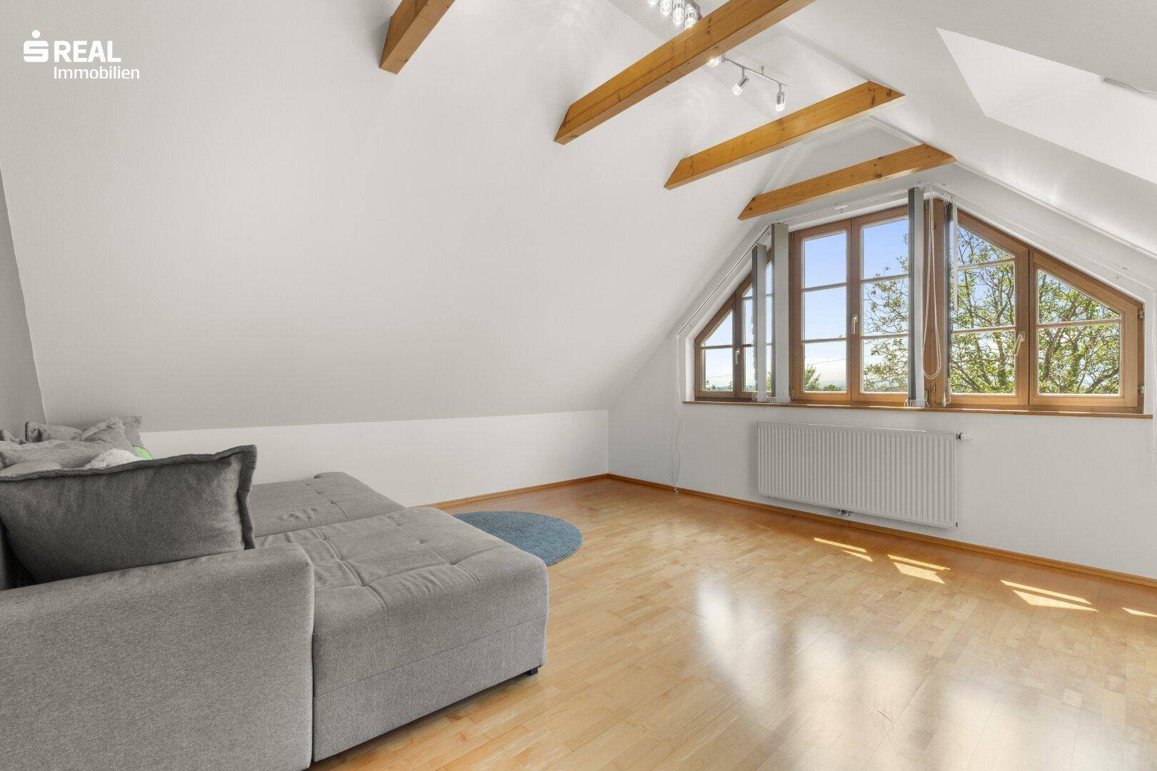 Gemütliches Wohnzimmer im Dachgeschoss mit Holzbalken, Dachschrägen und großen Fenstern.