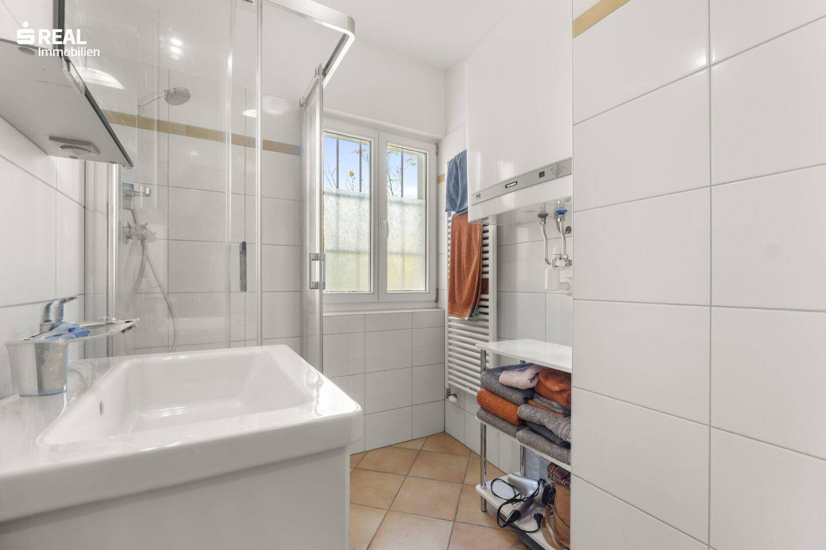 Kleines Badezimmer mit Dusche, Waschbecken und Fenster, ausgestattet mit Terrakotta-Fliesen.