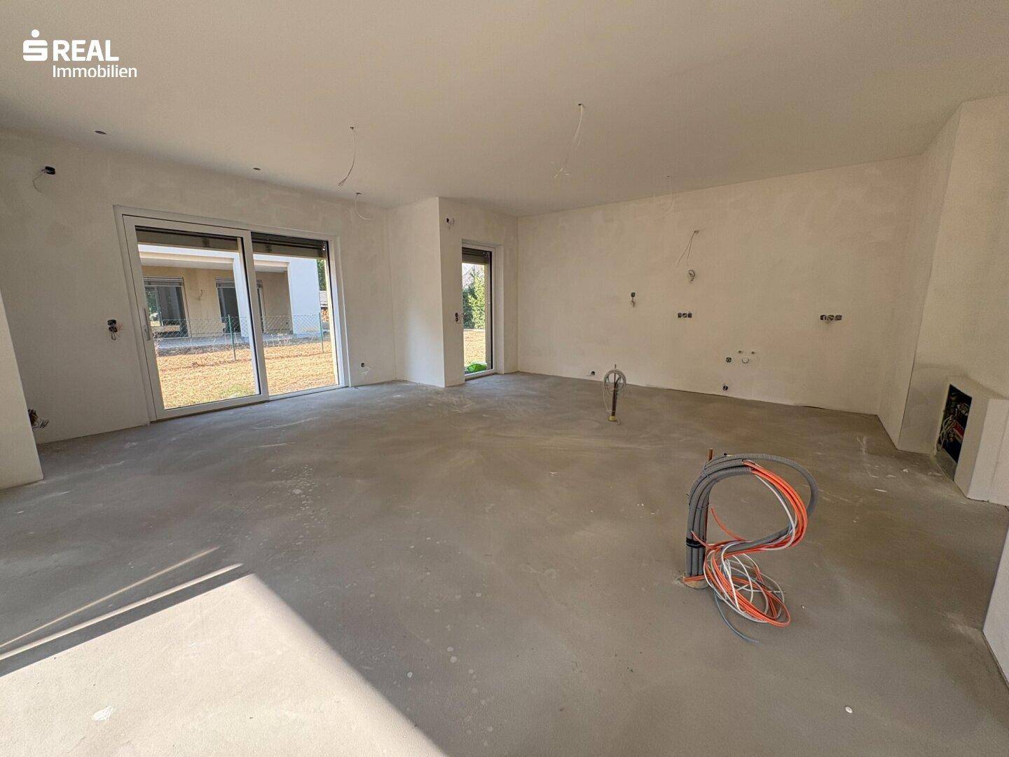 Geräumiger Wohnbereich im Rohbau mit großen Schiebetüren, die viel Tageslicht hereinlassen und zum Garten führen.