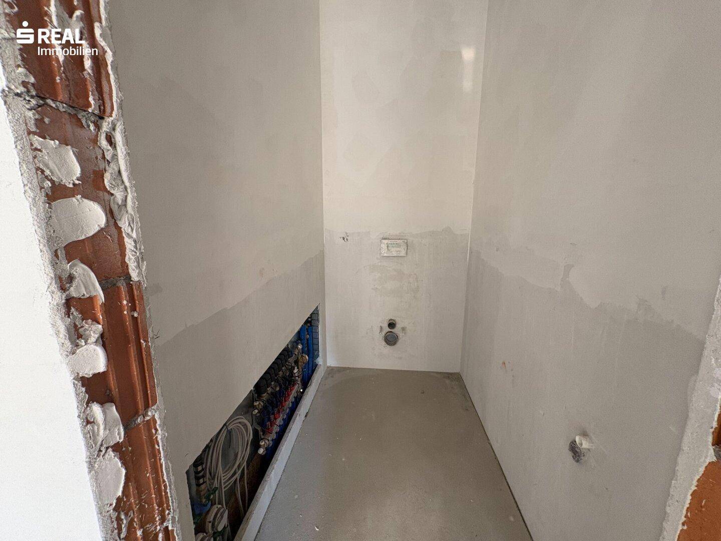 Kleiner Raum im Rohbau, vorbereitet für ein Gäste-WC mit sichtbaren Wasseranschlüssen und Abfluss.