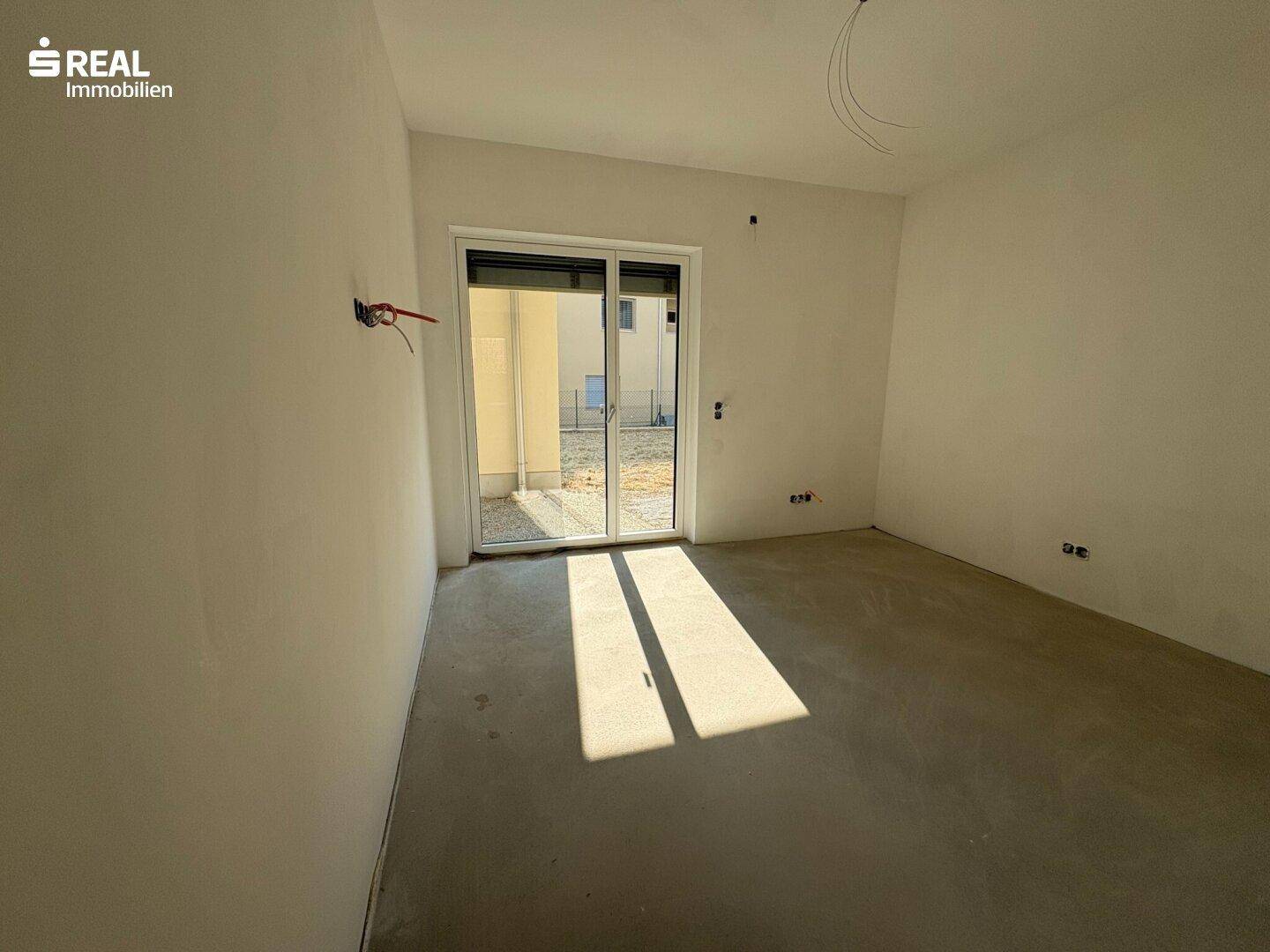 Heller Raum im Rohbau mit einer Schiebetür, die Zugang zum Außenbereich bietet und Sonnenlicht auf den Boden wirft.