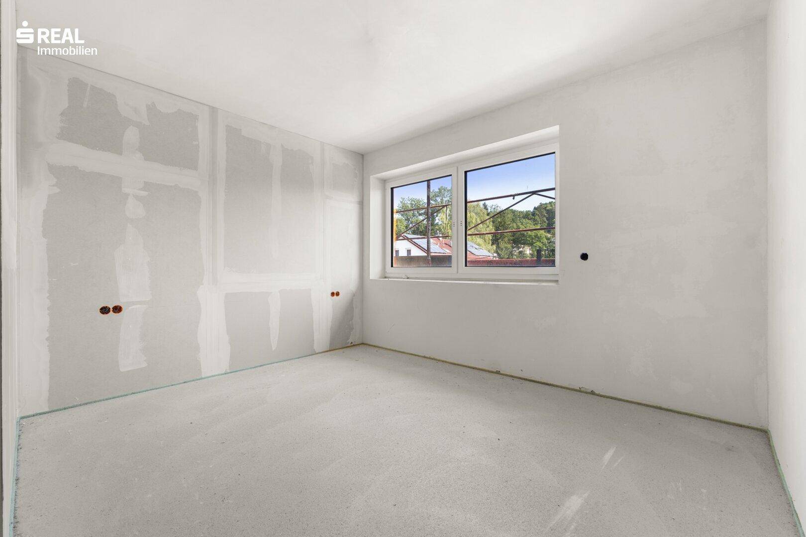 Unfertiges Zimmer im Rohbau mit einem Fenster, das einen Blick ins Grüne bietet.