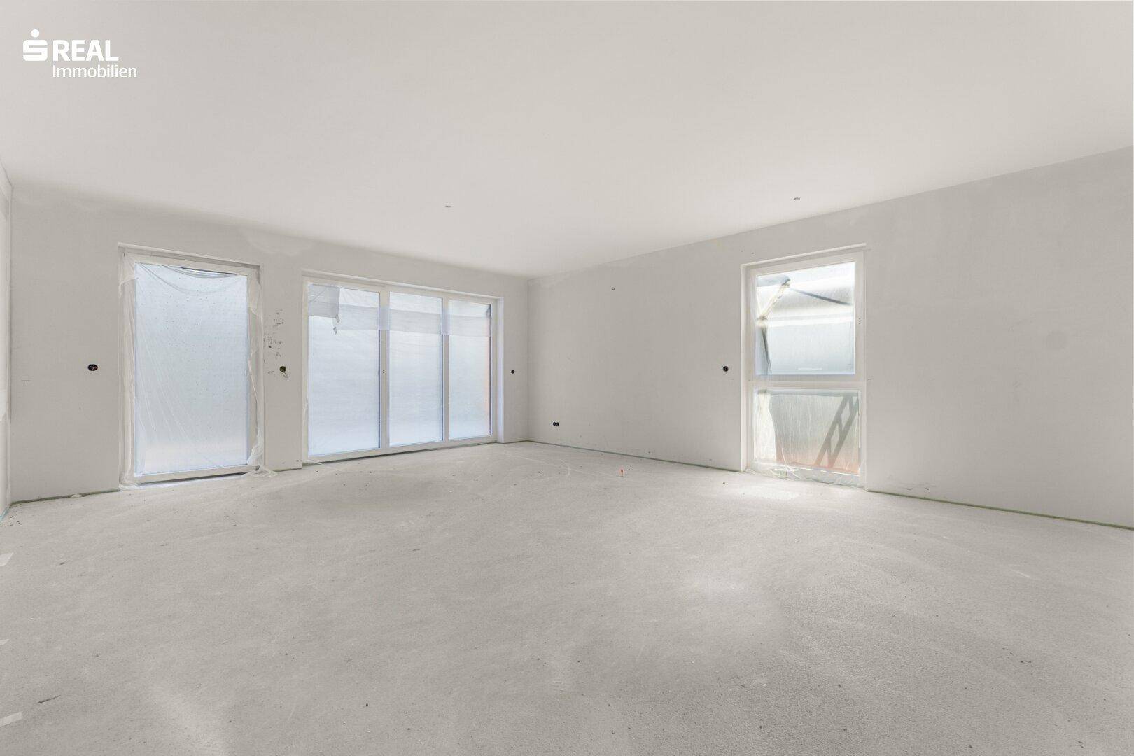 Unfertiger Wohnraum im Rohbau mit großen Fensterfronten, die viel natürliches Licht hereinlassen.
