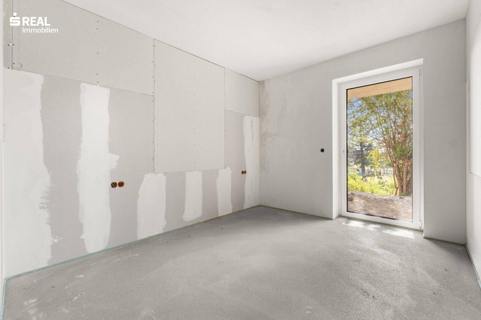 Rohbau eines Zimmers mit Trockenbauwänden, Betonboden und einem Fenster mit Blick ins Grüne.