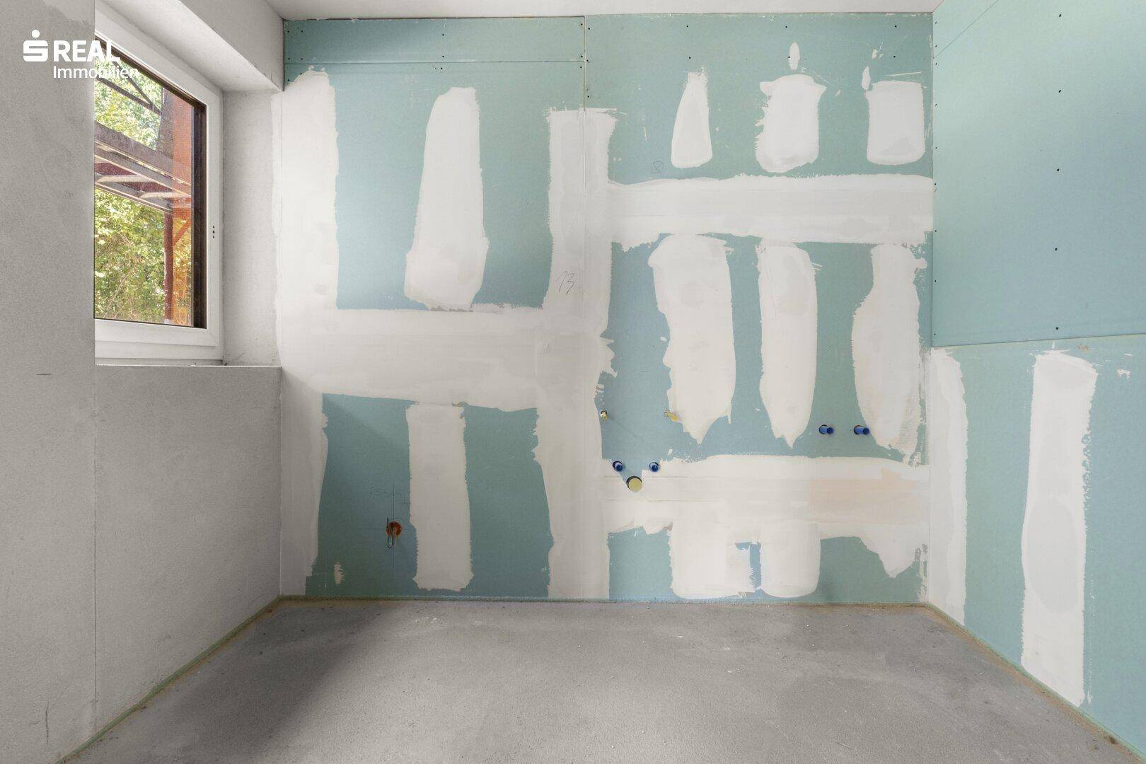 Unfertiges Badezimmer mit blauen Trockenbauwänden, sichtbaren Sanitäranschlüssen und einem kleinen Fenster.