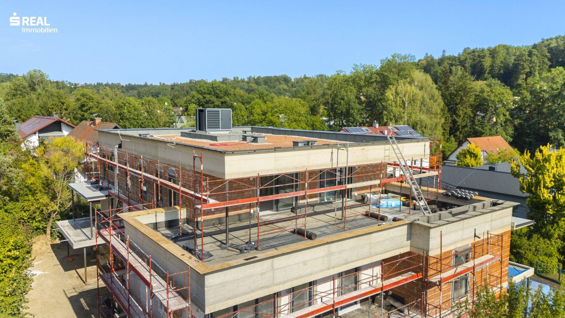Draufsicht auf das im Bau befindliche Gebäude mit Gerüst und Flachdach, umgeben von Bäumen.