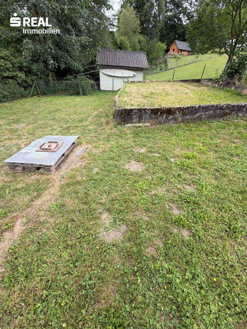Gartenbereich mit einer Feuerstelle und einem kleinen Schuppen im Hintergrund, umgeben von Grün.