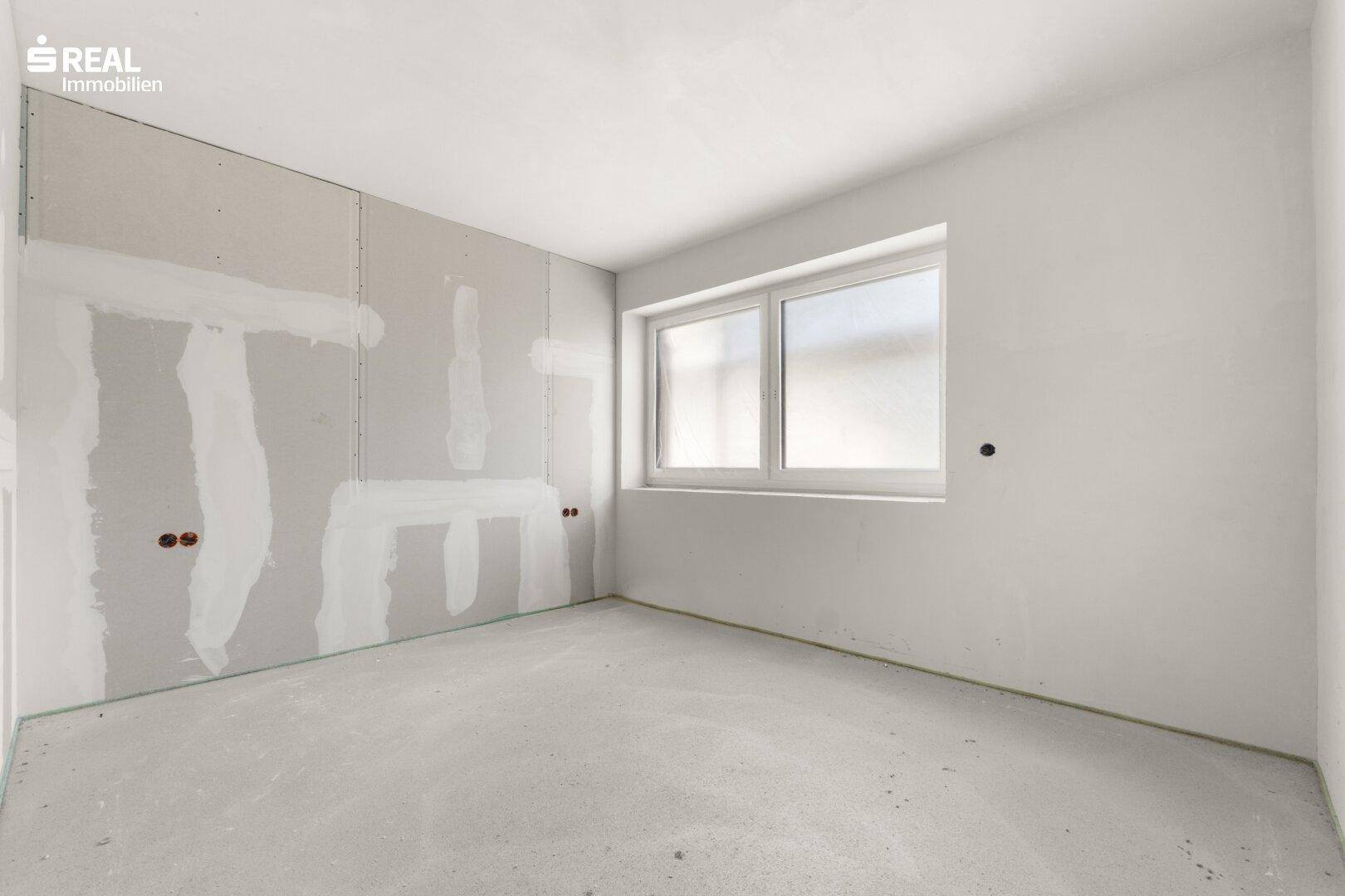 Unfertiger Raum mit Trockenbauwänden, Betonboden und einem Fenster, das auf die Baustelle blickt.