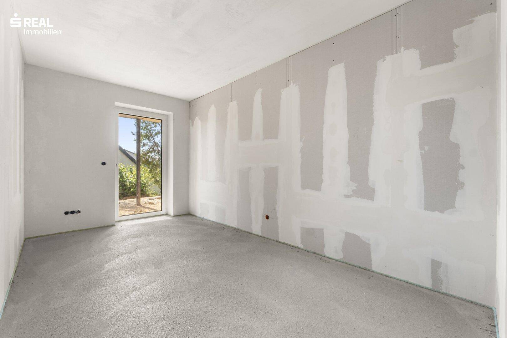 Rohbau eines Zimmers mit unverputzten Wänden, Betonboden und einem Fenster, das Tageslicht hereinlässt.