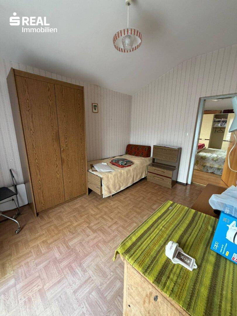 Schlafzimmer mit Einzelbett, Holzschrank und gemustertem Boden, mit Blick in den Nebenraum.