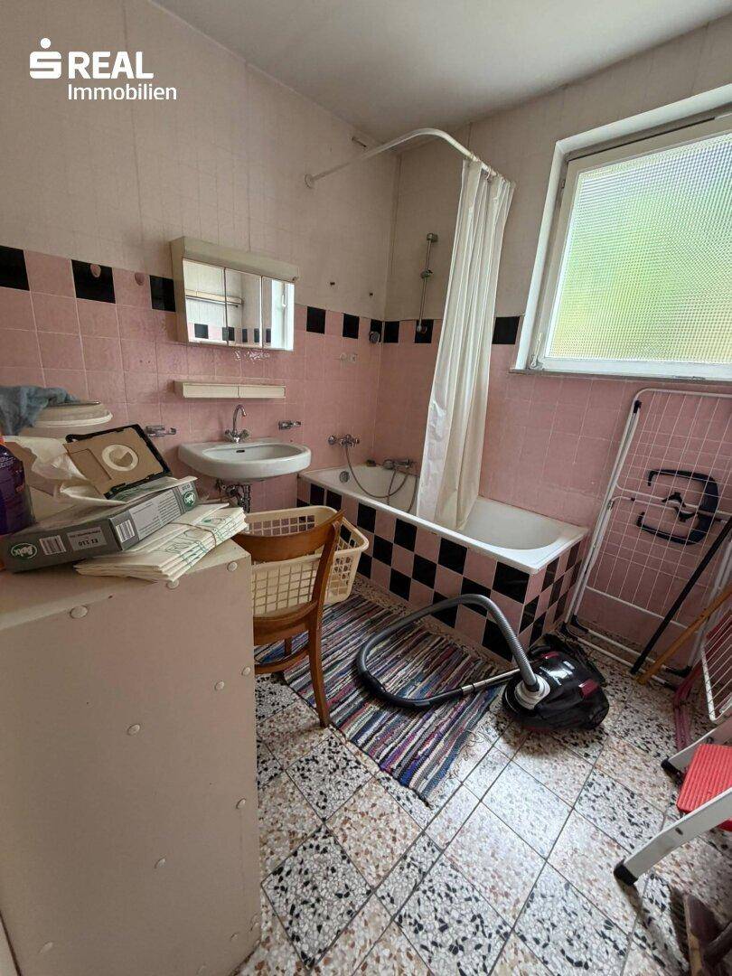 Badezimmer mit Badewanne, Waschbecken und rosa-schwarz gefliesten Wänden, renovierungsbedürftig.