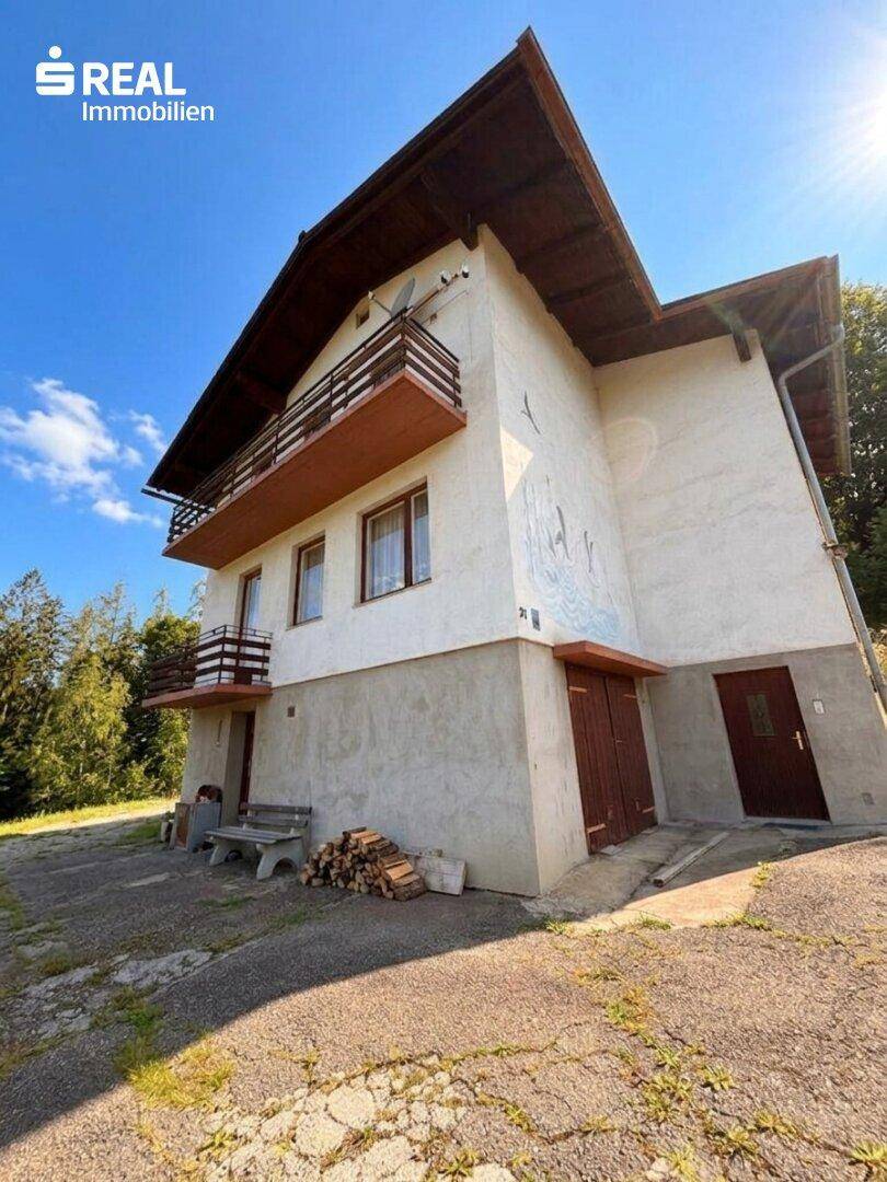 Ansicht des Hauses mit Balkonen, Holzlager und Zufahrt, umgeben von viel Grün.