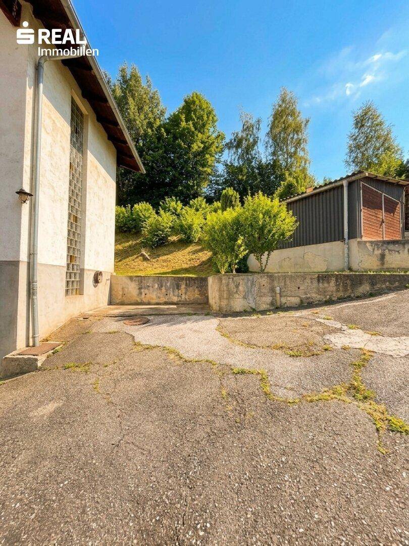 Außenbereich mit asphaltierter Fläche, einem Schuppen und viel Grün im Hintergrund.