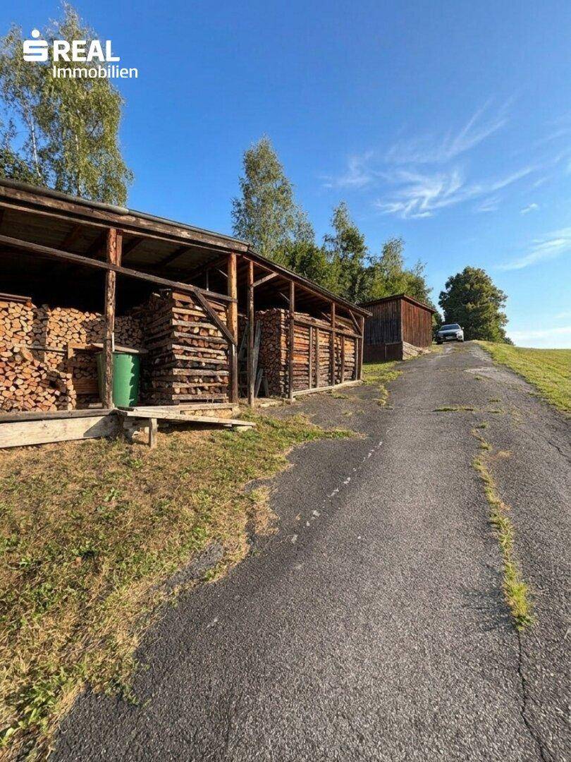 Großer Holzschuppen mit gestapeltem Brennholz entlang einer ländlichen Zufahrtsstraße.