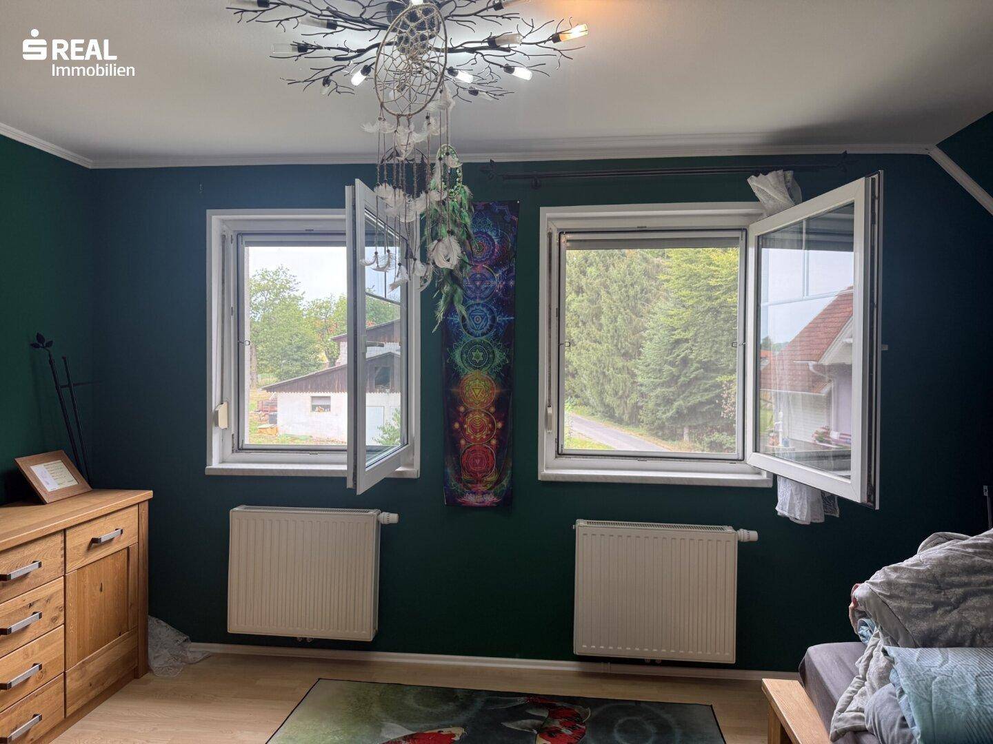 Schlafzimmer mit dunkelgrünen Wänden, Holzkommode und Blick ins Grüne.