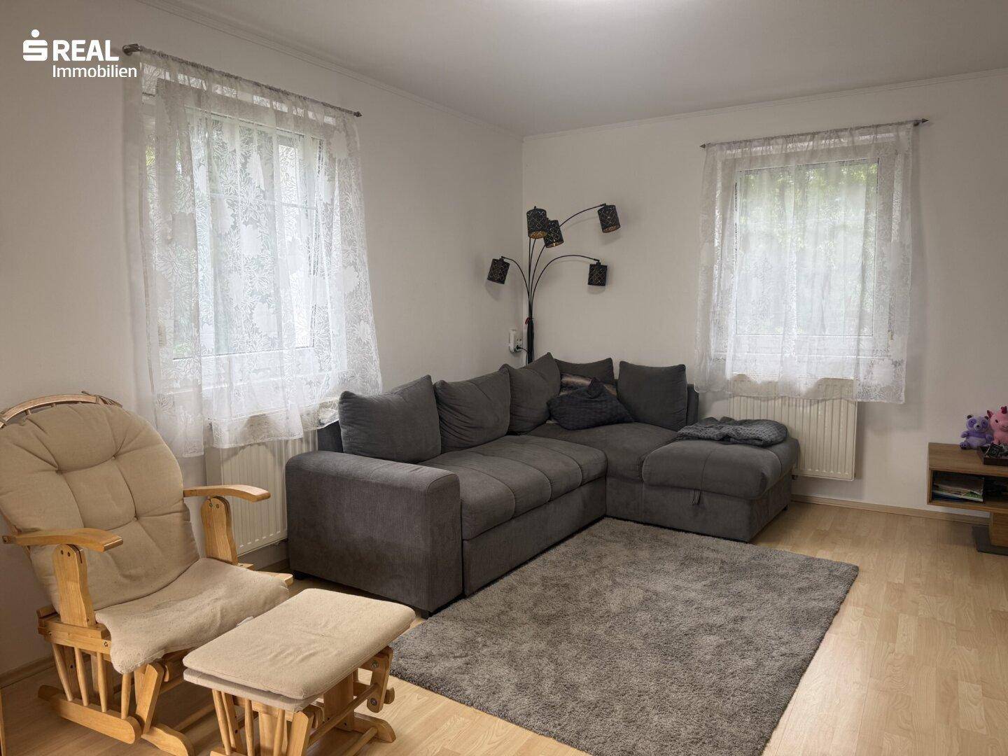 Helles Wohnzimmer mit großem Ecksofa, Schaukelstuhl und zwei Fenstern.