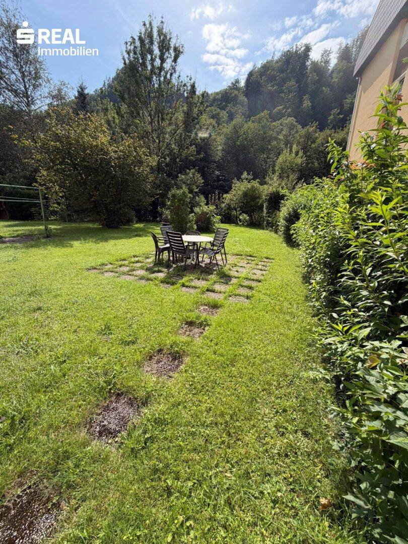 Gemütlicher Sitzbereich im Garten mit Steinfliesen und umgebender grüner Bepflanzung.