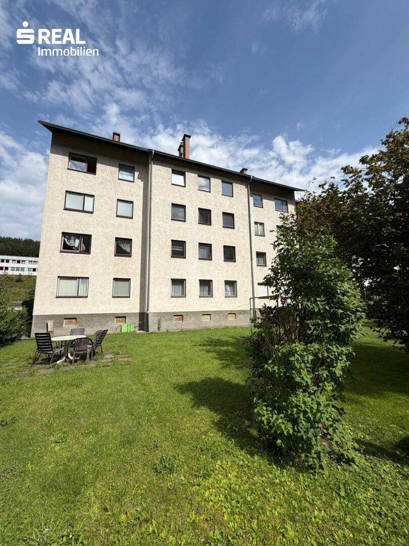 Rückansicht des Mehrfamilienhauses mit großem Rasenbereich und Bäumen im Hintergrund.