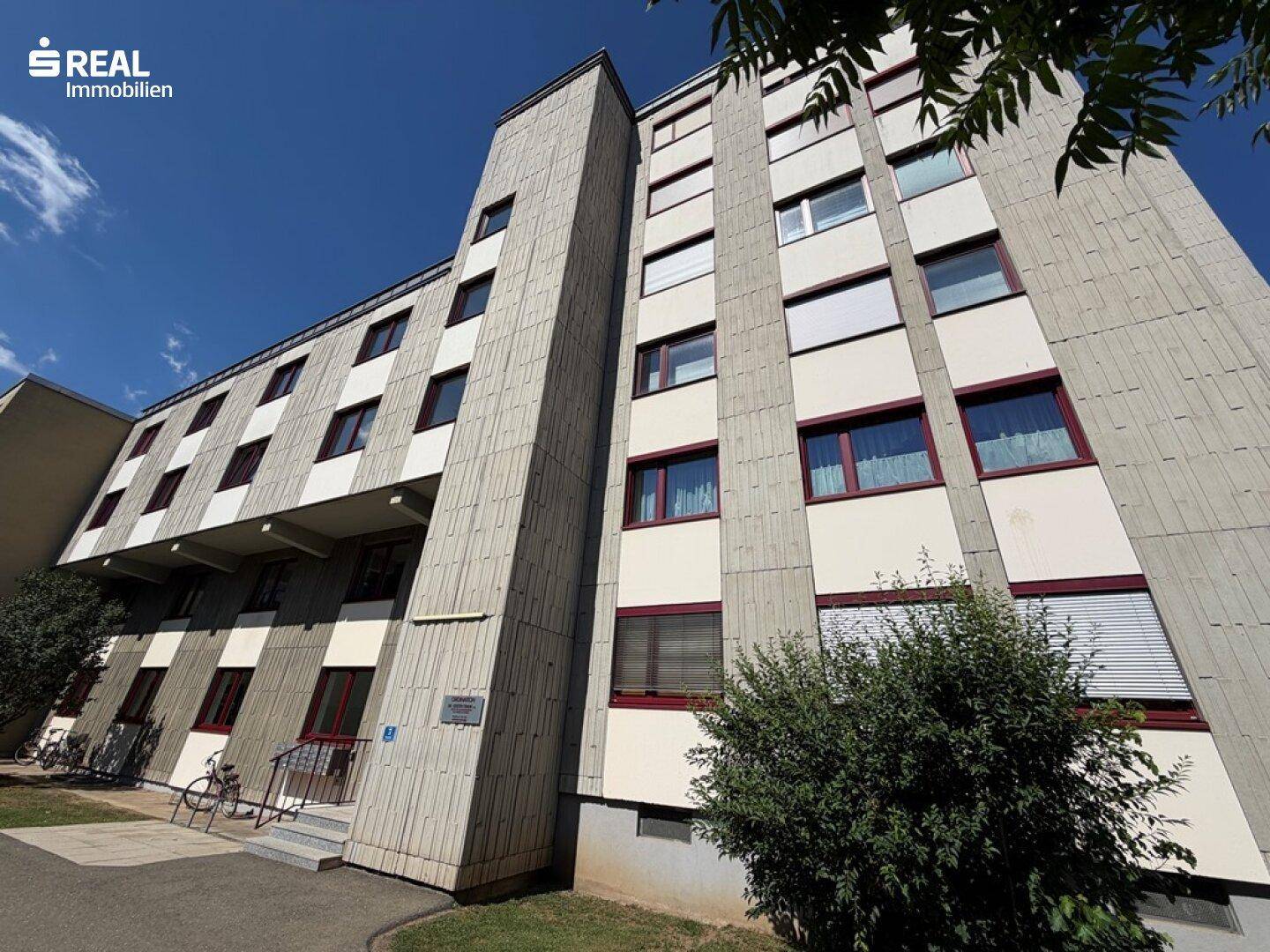 Detailansicht der Betonfassade eines Mehrfamilienhauses mit roten Fensterrahmen und Jalousien.