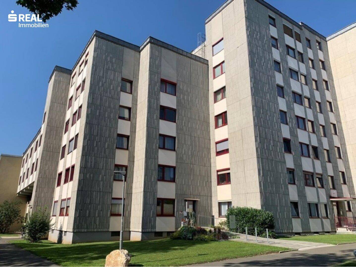 Weitläufige Ansicht eines modernen Mehrfamilienhauses mit Betonfassade und roten Akzenten in sonniger Umgebung.