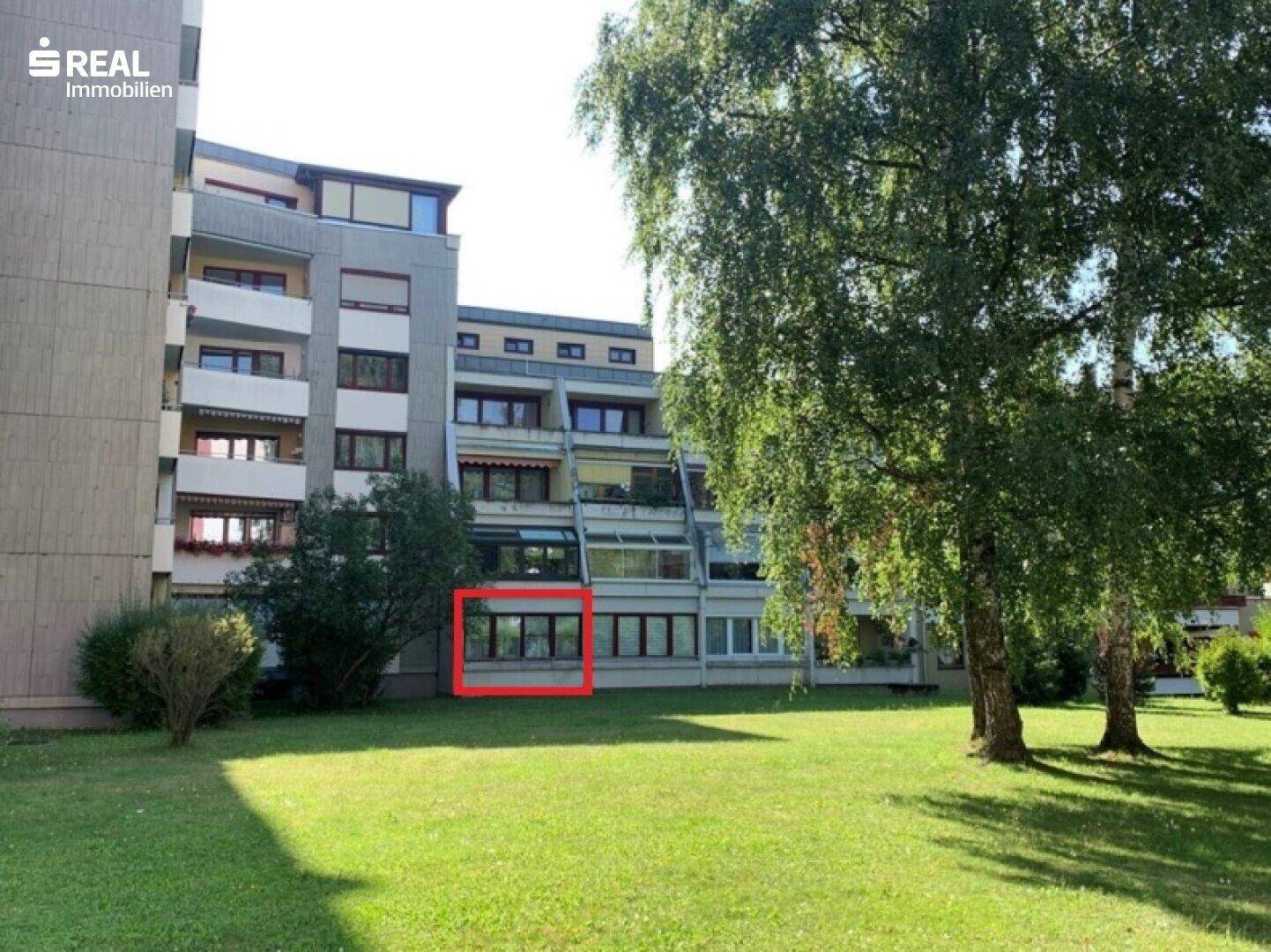 Grüne Rasenfläche vor einem Mehrfamilienhaus mit Balkonen und großen Fenstern, umgeben von Bäumen.