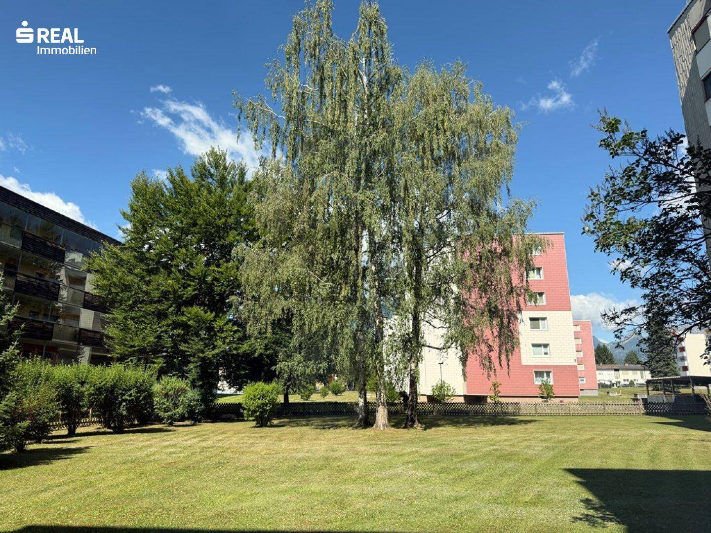Weitläufige Grünfläche mit hohen Bäumen und weiteren Wohngebäuden im Hintergrund unter blauem Himmel.