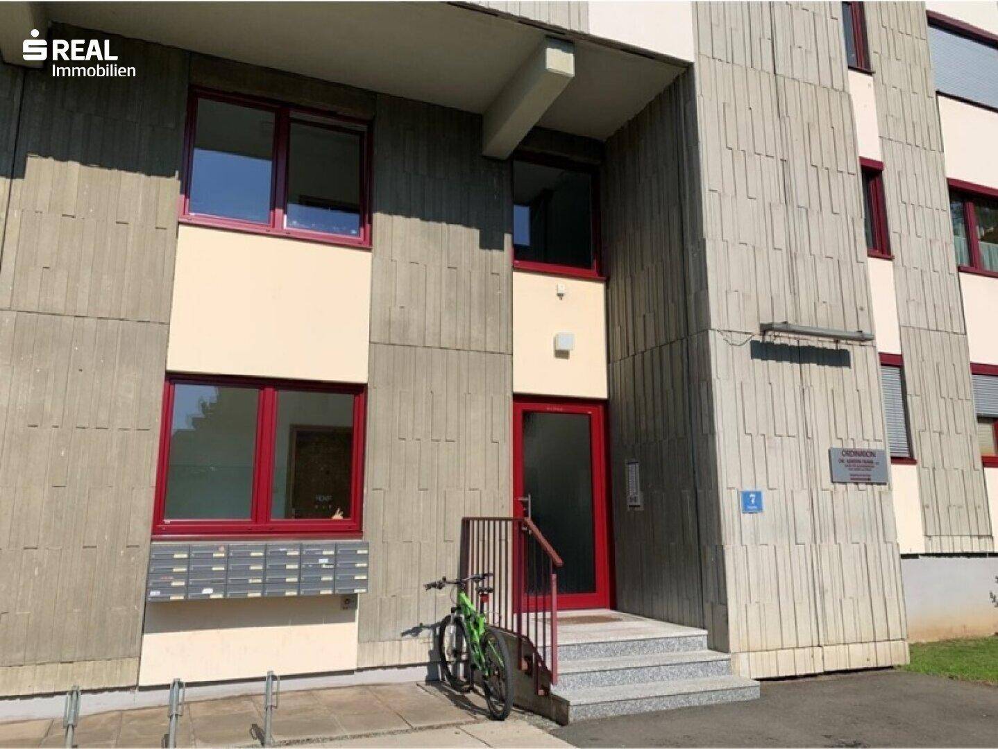 Eingangsbereich eines Mehrfamilienhauses mit roter Tür, Briefkästen und Fahrradabstellplatz.
