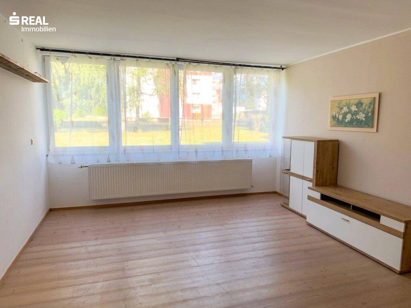 Heller Wohnbereich mit Holzboden, Heizkörper und großen Fenstern mit Blick ins Grüne.