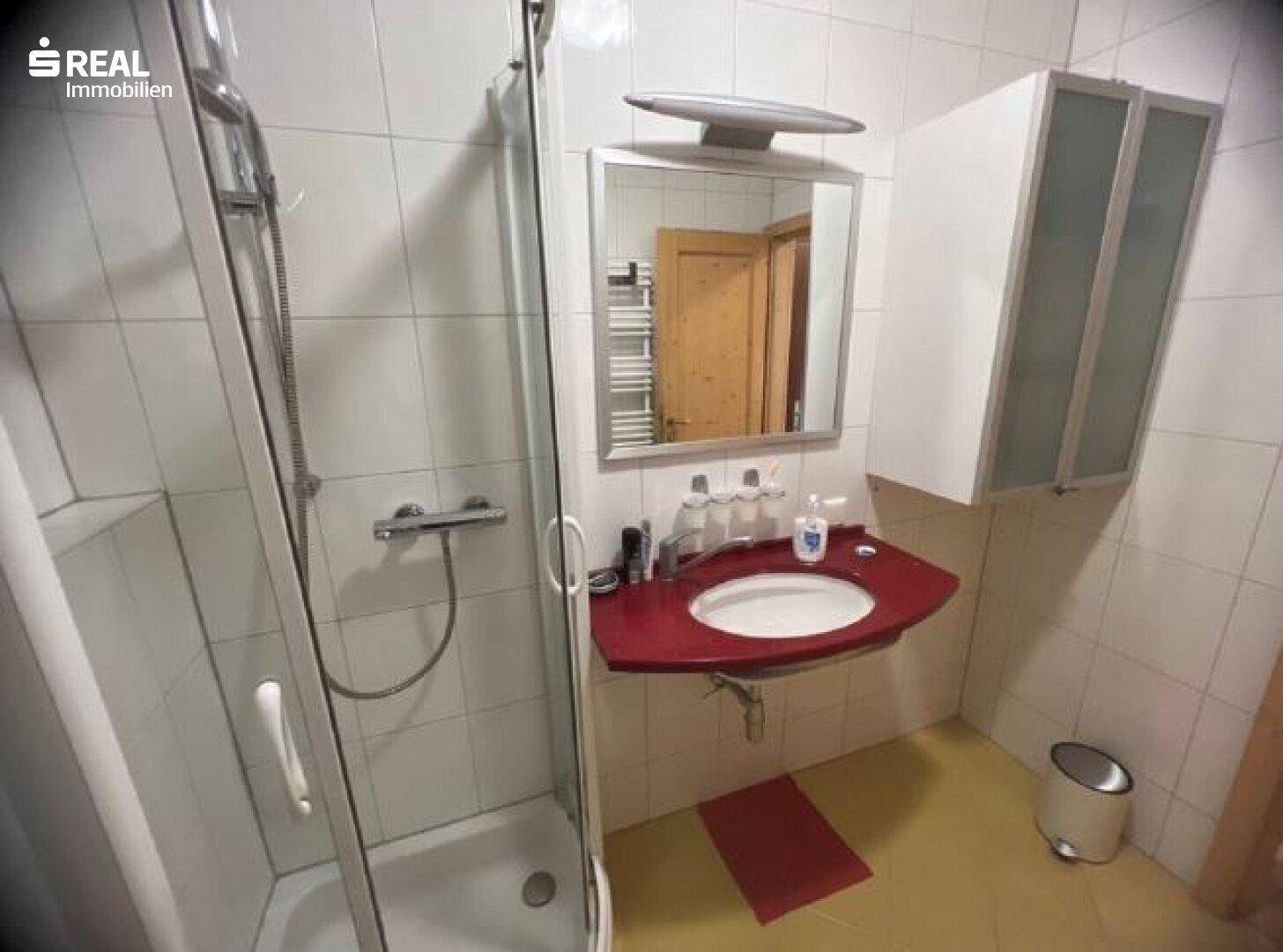 Helles Badezimmer mit Duschkabine, Waschbecken mit roter Ablage und Spiegelschrank.