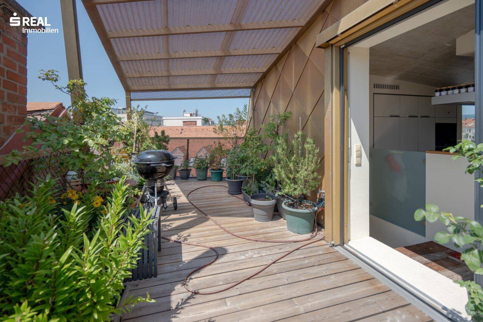 Breite Holzterrasse mit Grill, vielen Pflanzen und Zugang zur Küche.