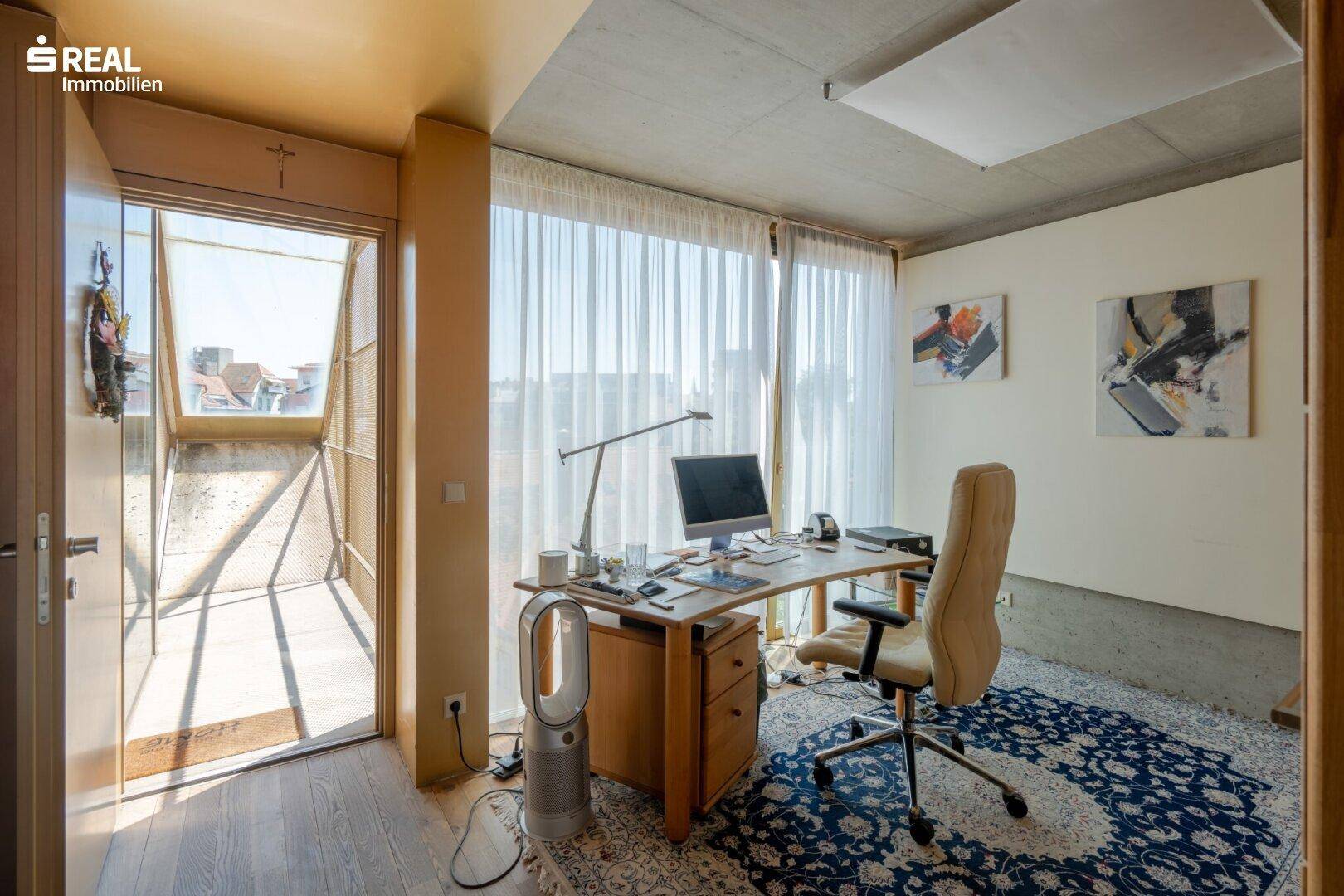 Helles Arbeitszimmer mit großem Schreibtisch, Teppich und Zugang zu einem Balkon.