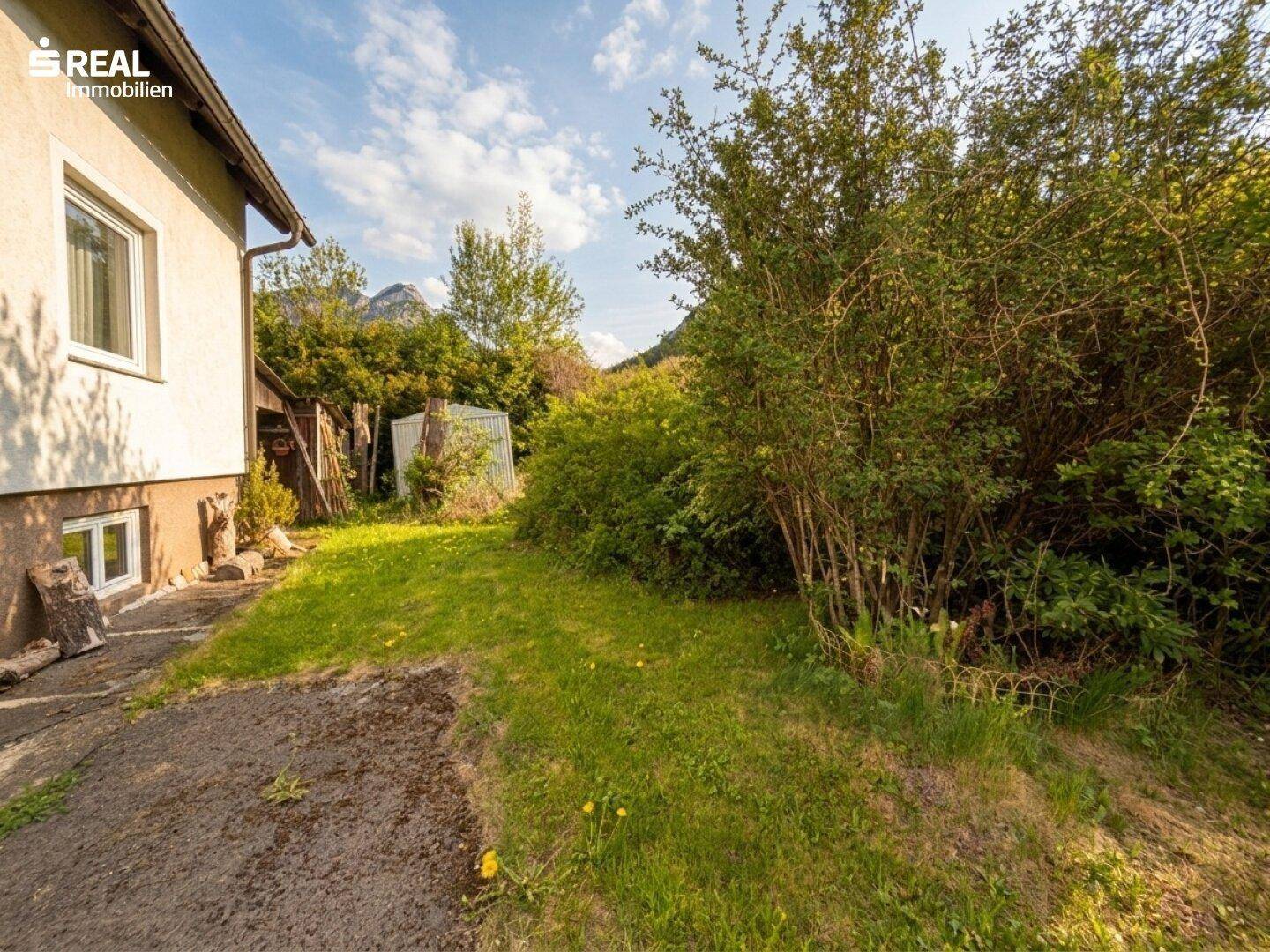 Grüner Gartenbereich neben dem Haus mit Sträuchern und Blick auf die bewaldeten Hügel.