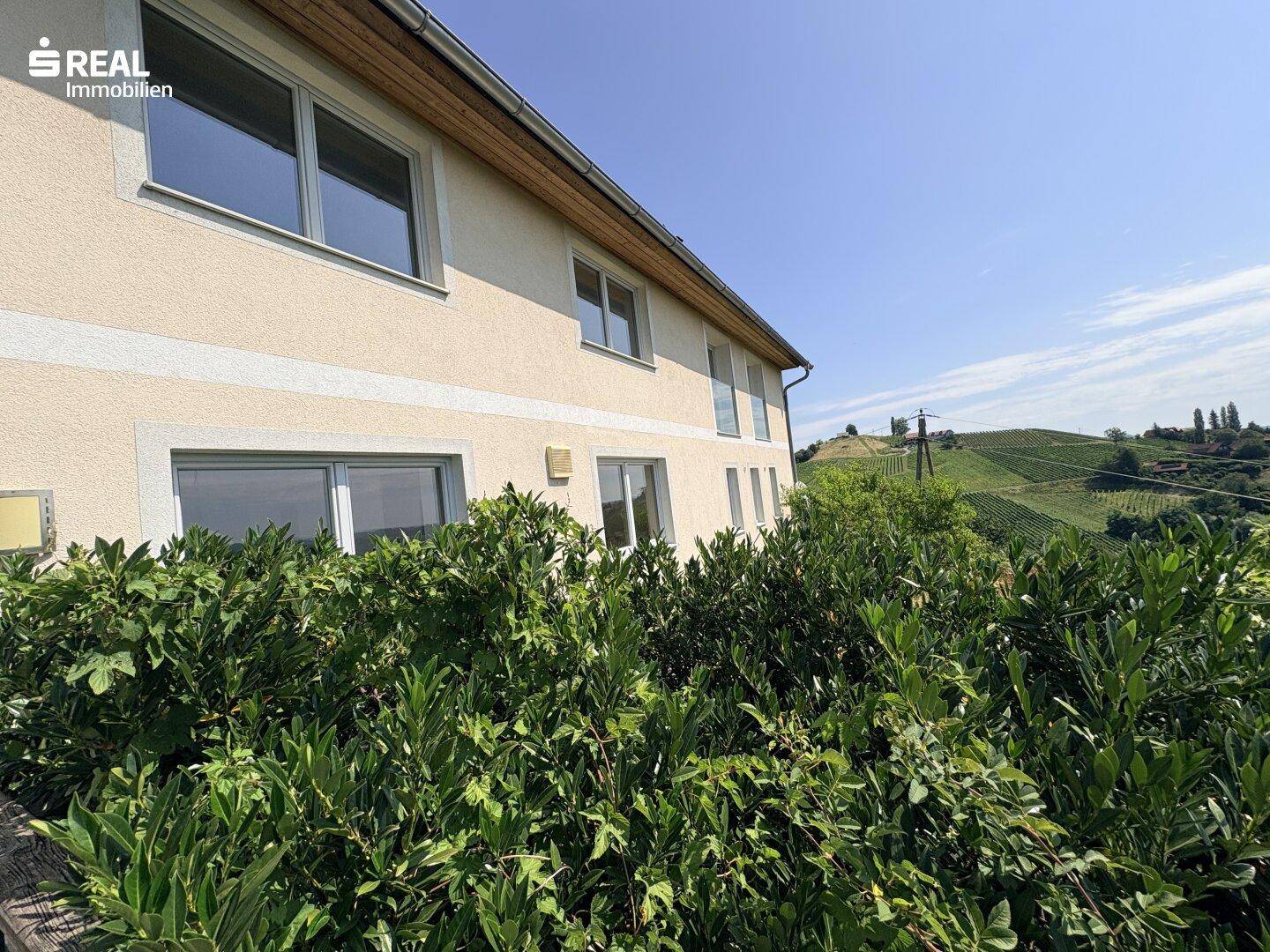 Seitenansicht des Hauses mit großen Fenstern und üppigen Büschen im Vordergrund, Blick auf Weinberge.