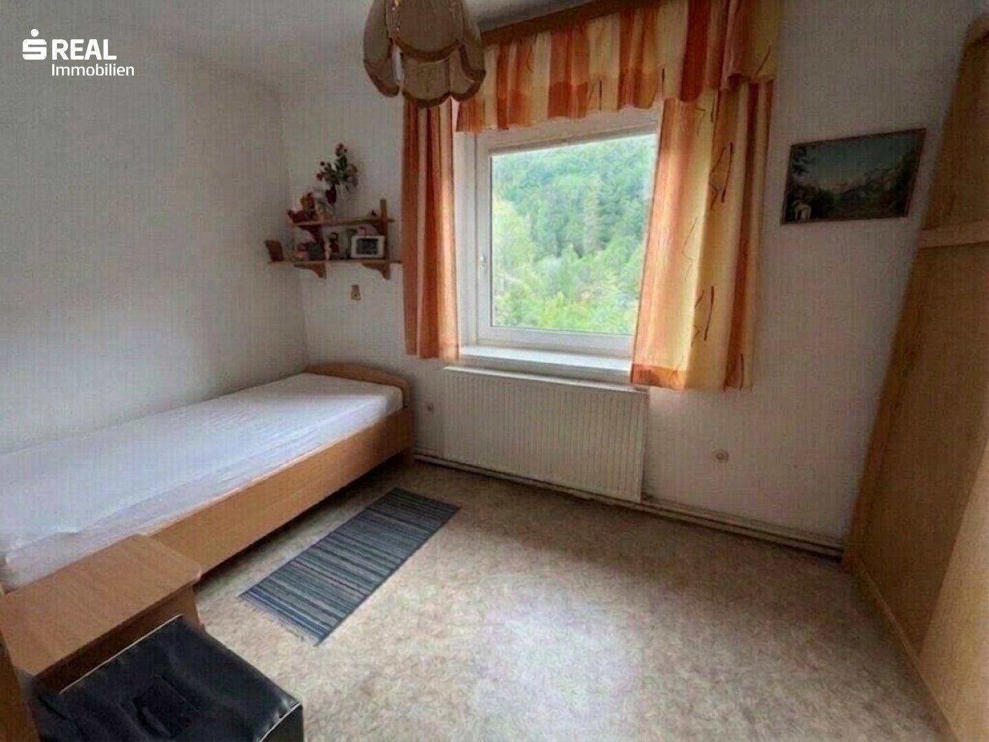Einfaches Schlafzimmer mit Einzelbett, Teppichboden und Fenster mit Blick ins Grüne.