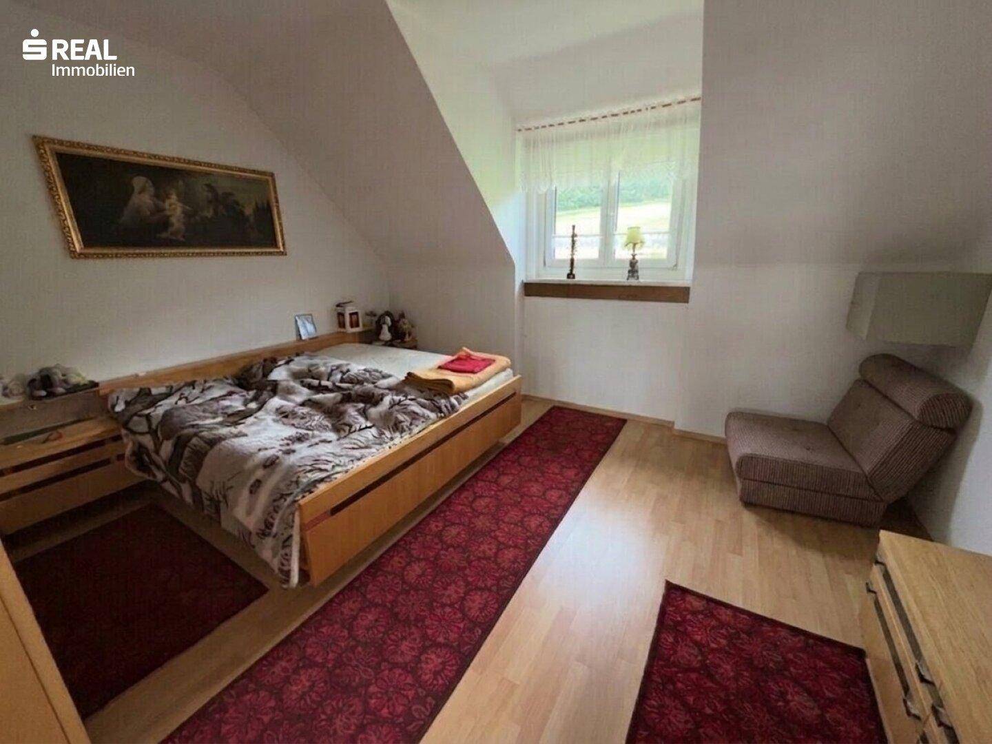 Schlafzimmer im Dachgeschoss mit Holzbett, Teppich und Fenster mit Blick ins Grüne.