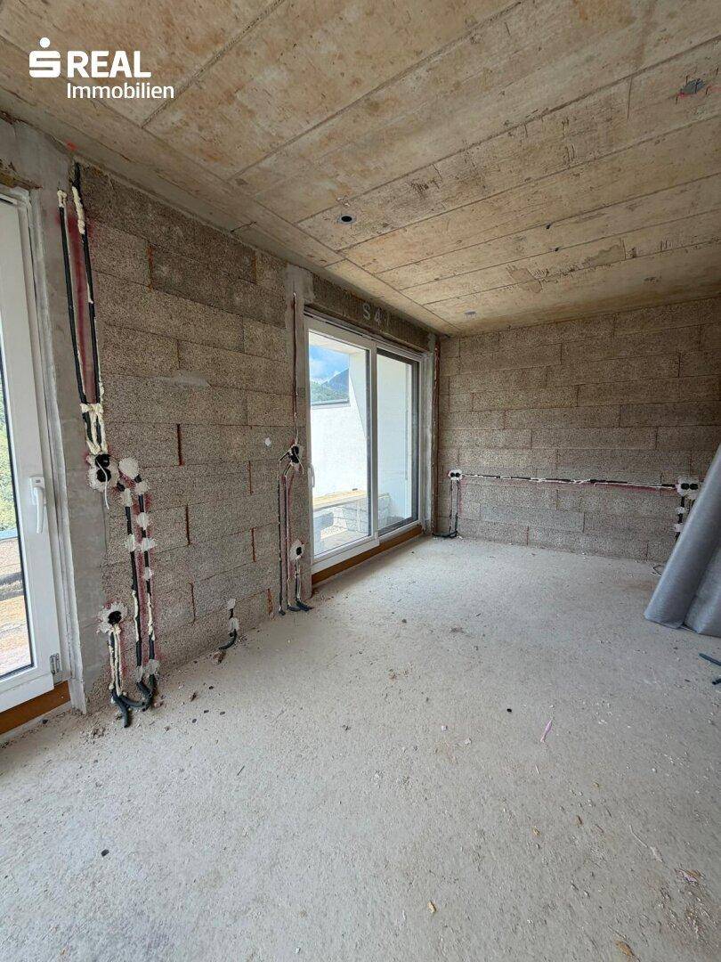 Rohbauzimmer mit unverputzten Wänden, großen Fenstern und vorbereiteten Elektroinstallationen.